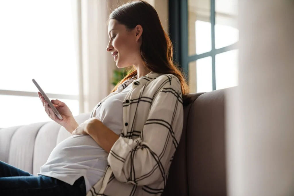 Happy charming pregnant woman using mobile phone while sitting on sofa in cozy room