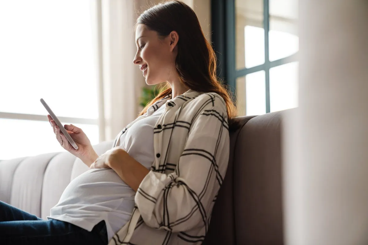Happy charming pregnant woman using mobile phone while sitting on sofa in cozy room